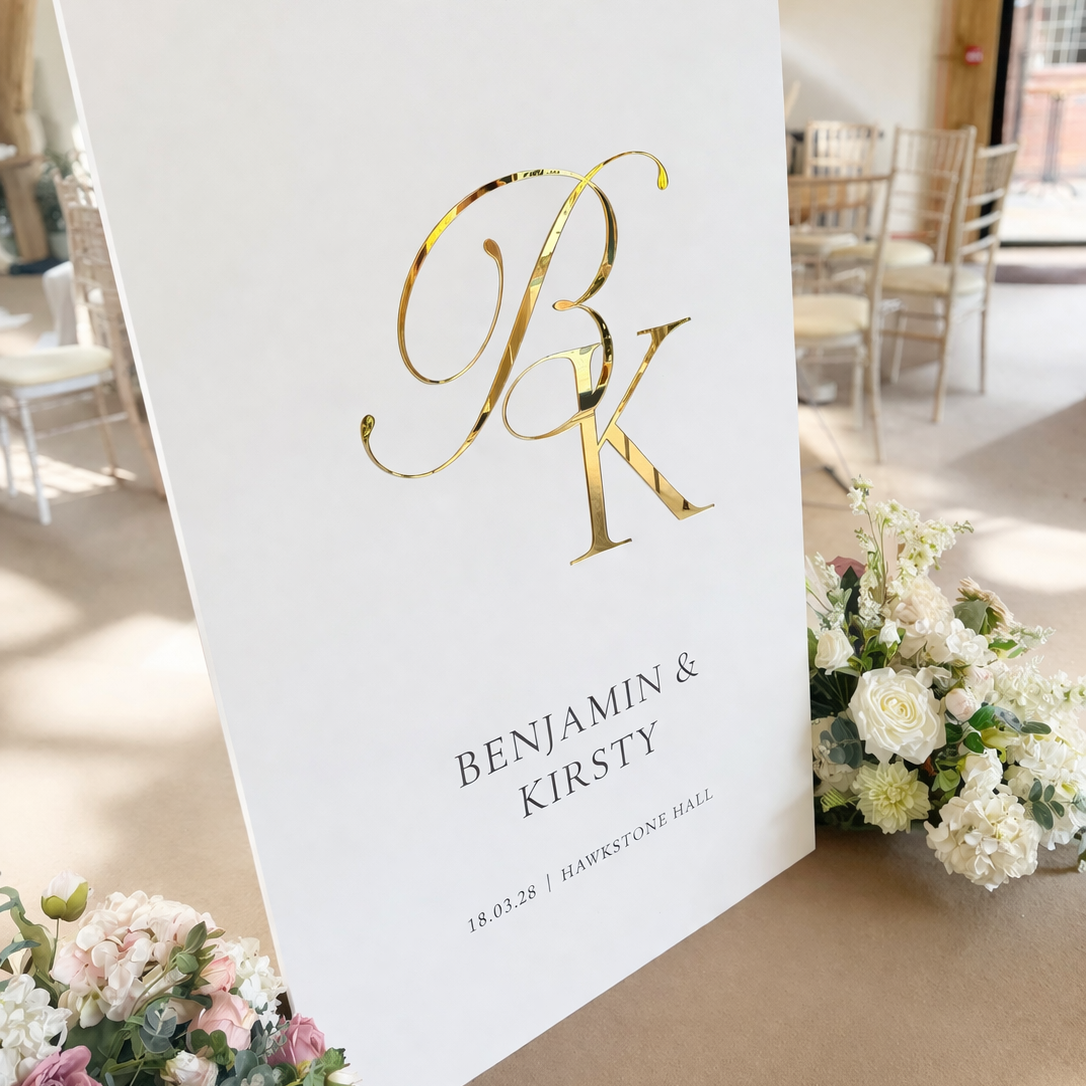 A white sign with the initials "BK" in gold lettering, displaying the names "BENJAMIN & KIRSTY" and the date "18.03.28" in black text. The sign is surrounded by a bouquet of white flowers and placed on a table.