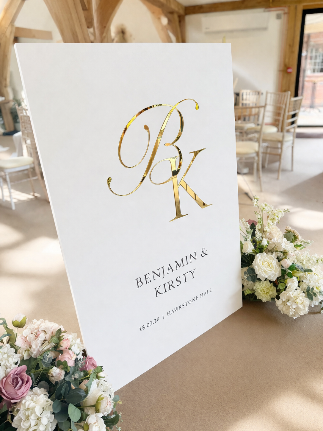A white sign with the initials "BK" in gold lettering, displaying the names "BENJAMIN & KIRSTY" and the date "18.03.28" in black text. The sign is surrounded by a bouquet of white flowers and placed on a table.