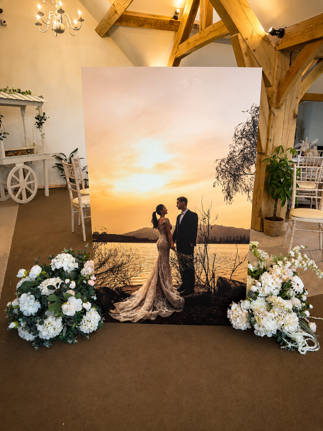A large wedding photograph of a couple standing on a beach at sunset, with a chandelier and floral arrangements in the background.