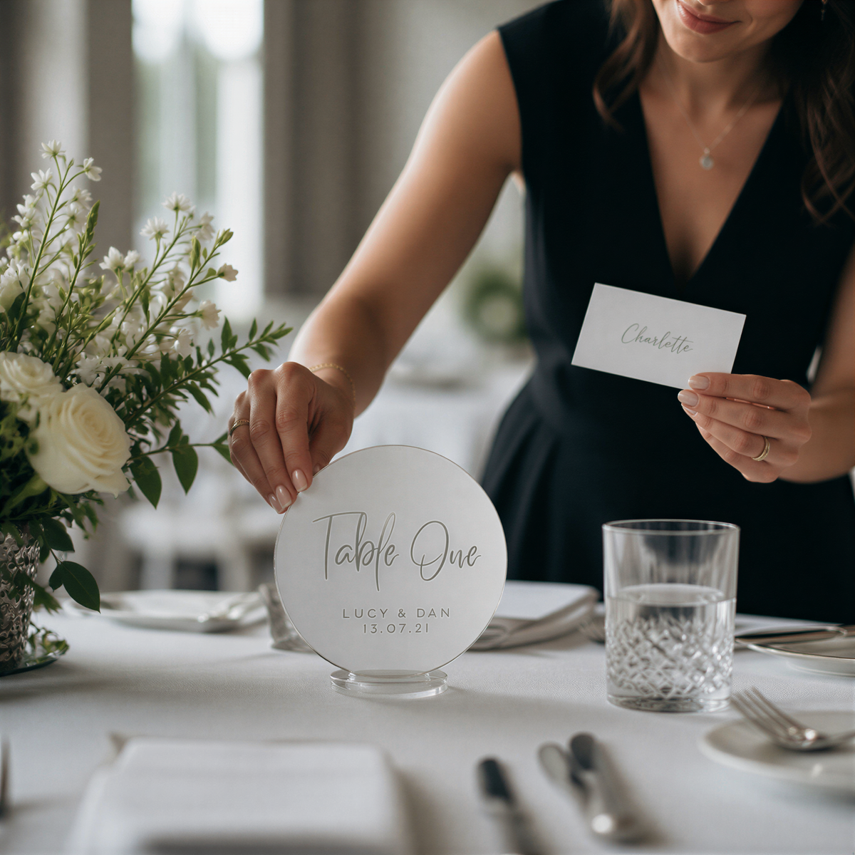 A woman is standing at a table with a table number sign, a glass of water, and a vase of white flowers.