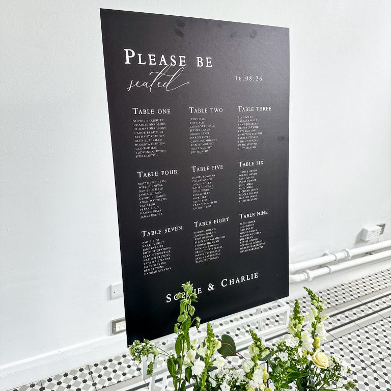 a wedding reception setup with a black and white colour scheme. There are two signs, one white and one black, both displaying the names of the couple, Sophie and Charlie. The white sign is positioned in front of the black sign, which is placed on a white stand. The stand also has a small floral arrangement on it.