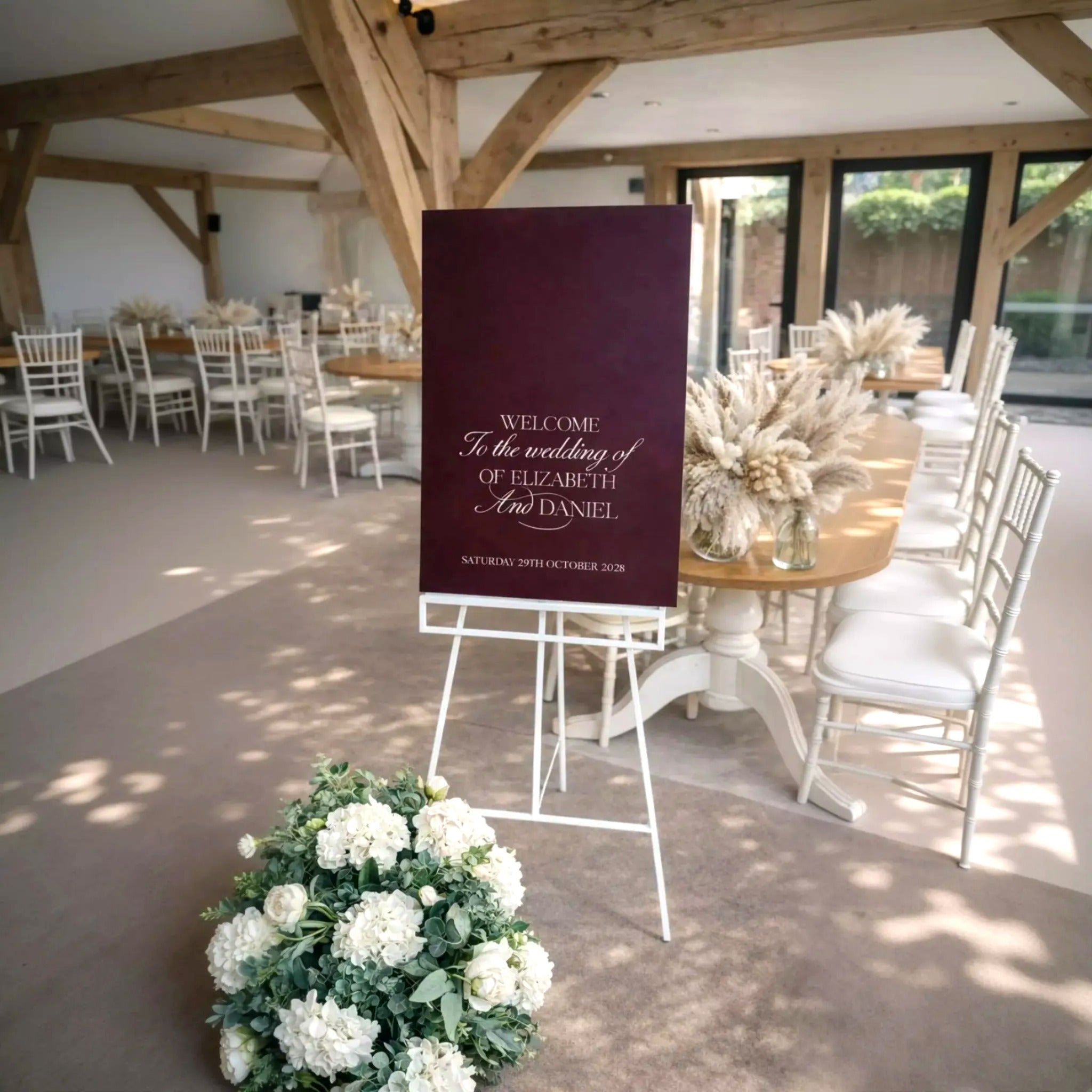 a wedding reception setup with a large sign welcoming guests to the wedding of Elizabeth and Daniel. The sign is placed in a room with tables and chairs, and there are potted plants and flowers decorating the space.