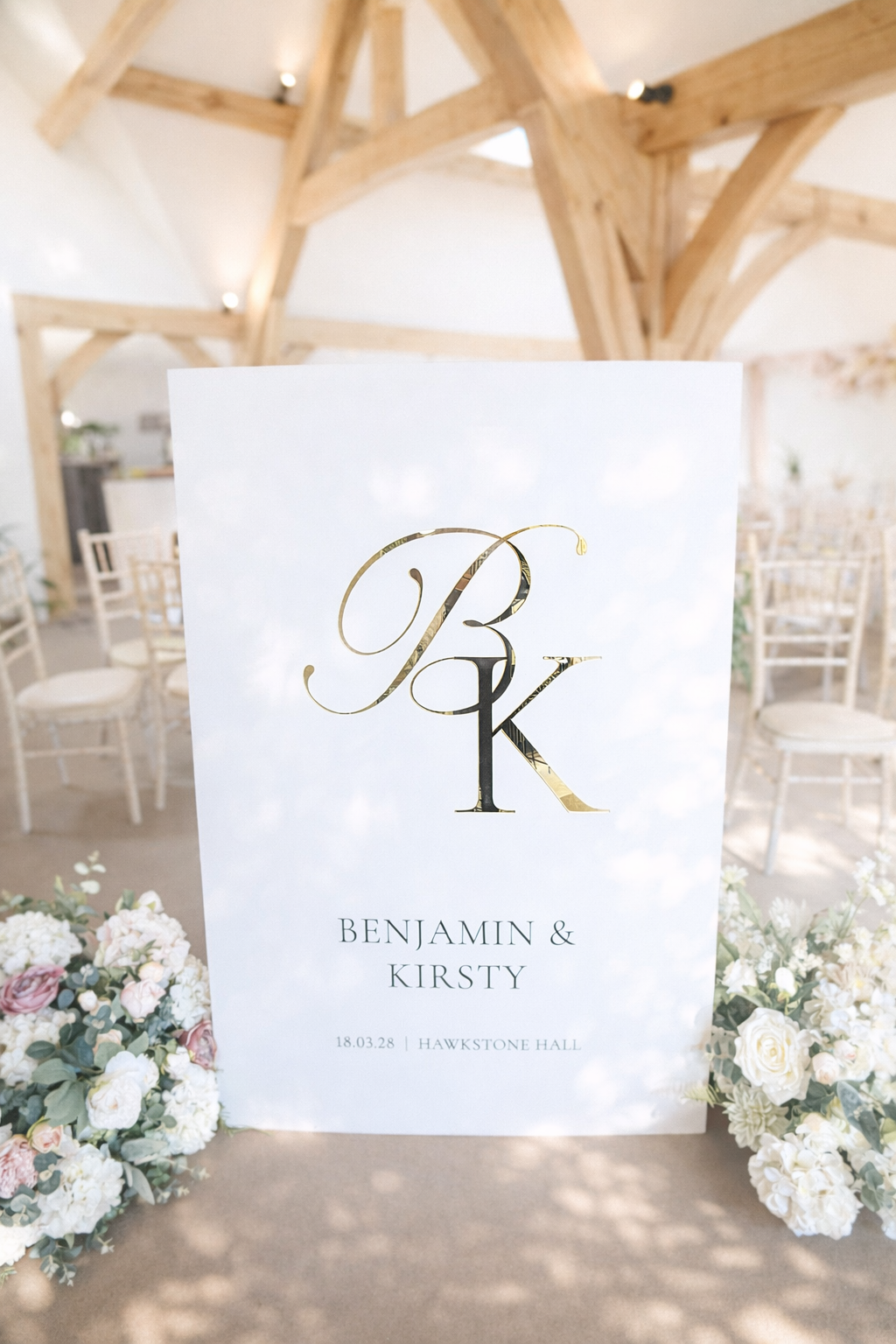 A white sign with the initials "BK" and the names "Benjamin & Kirsty" is displayed in a room with wooden beams and white chairs.