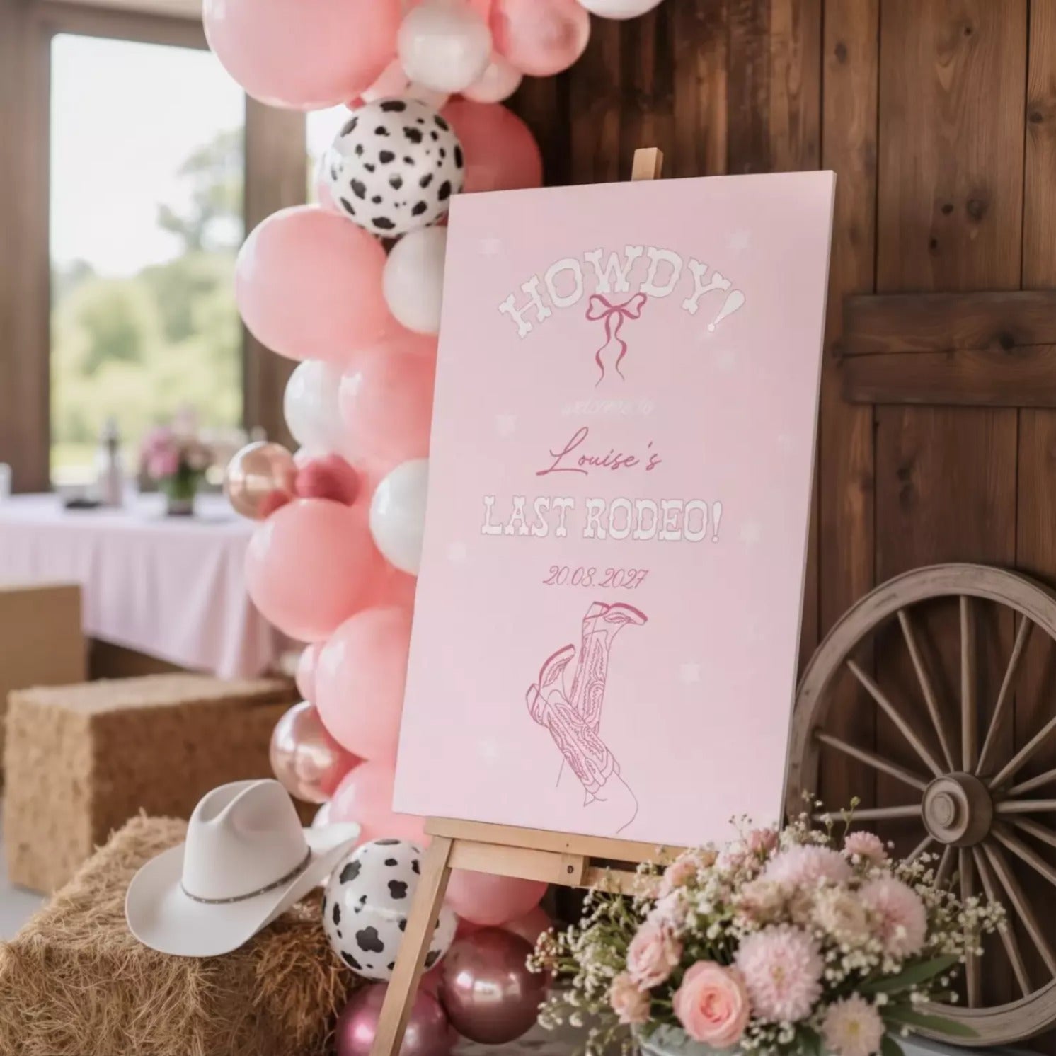 a pink sign with white text and a graphic of a cowboy hat, surrounded by pink and white balloons, a bouquet of pink flowers, and a wooden wagon wheel.