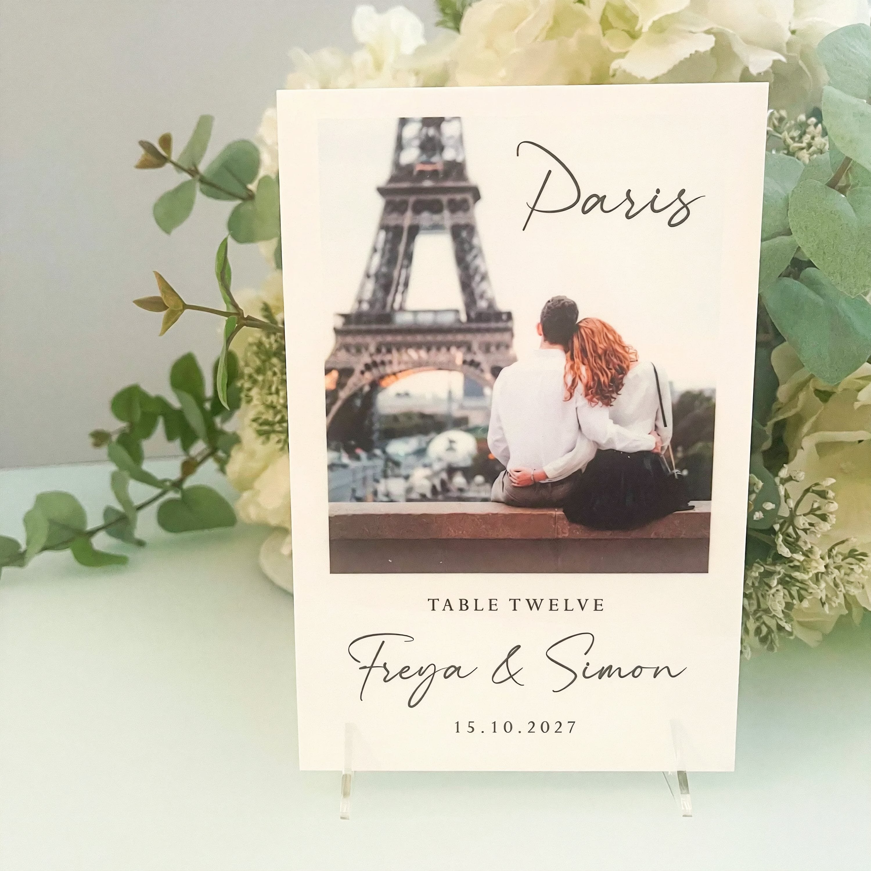 A wedding invitation card featuring a photo of a couple sitting on a bench in front of the Eiffel Tower, with the text "Paris" and "Table Twelve" printed on it.