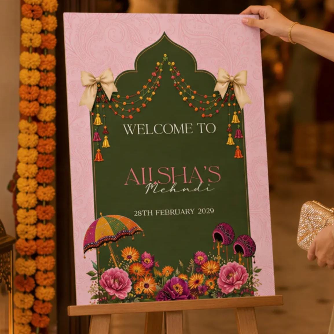 A woman in a green dress is holding a sign that reads "Welcome to Alishu's" in front of a pink and green sign with flowers and a decorative arch.