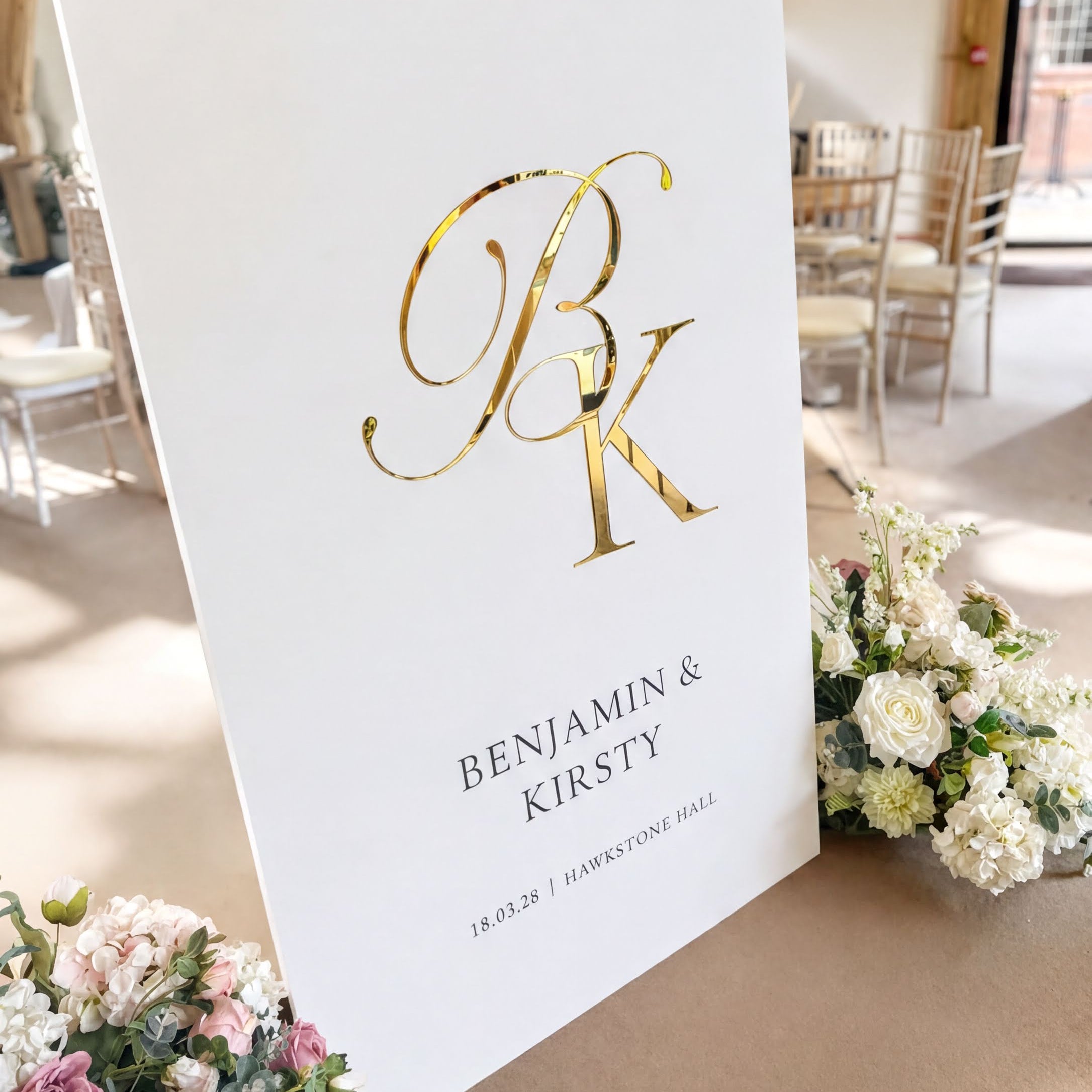 A white sign with the initials "BK" in gold lettering, displaying the names "BENJAMIN & KIRSTY" and the date "18.03.28" in black text. The sign is surrounded by a bouquet of white flowers and placed on a table.