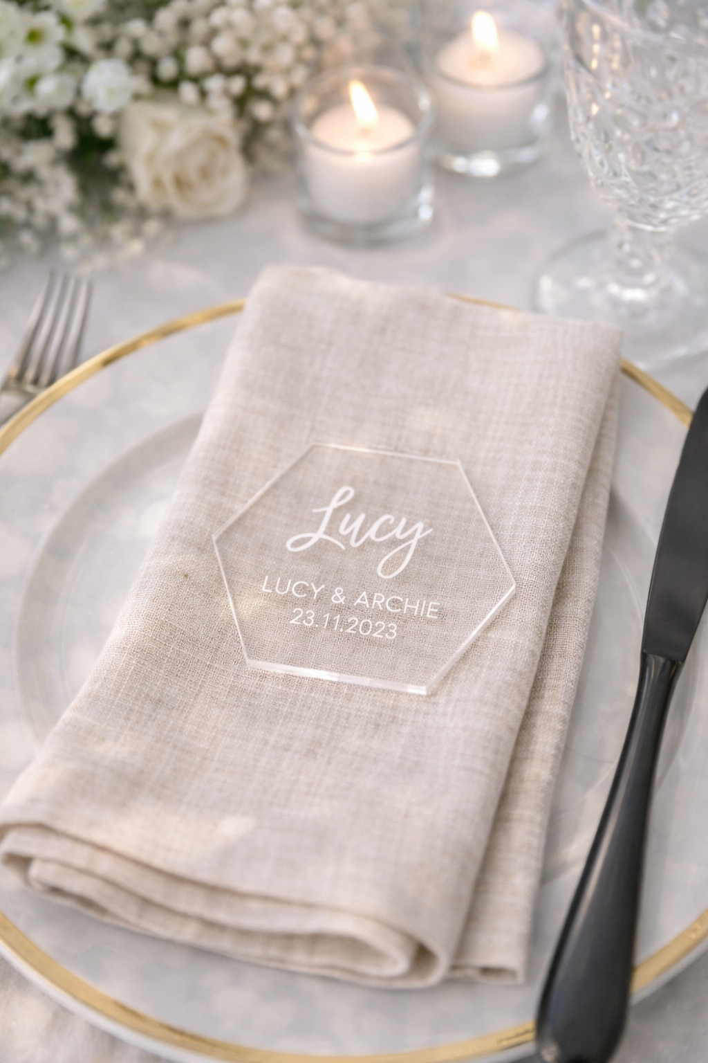 A table setting with a white napkin, a glass of water, and a candle, with a clear hexagonal placemat displaying the name 'Lucy & Archie' and the date '23.11.2023'.