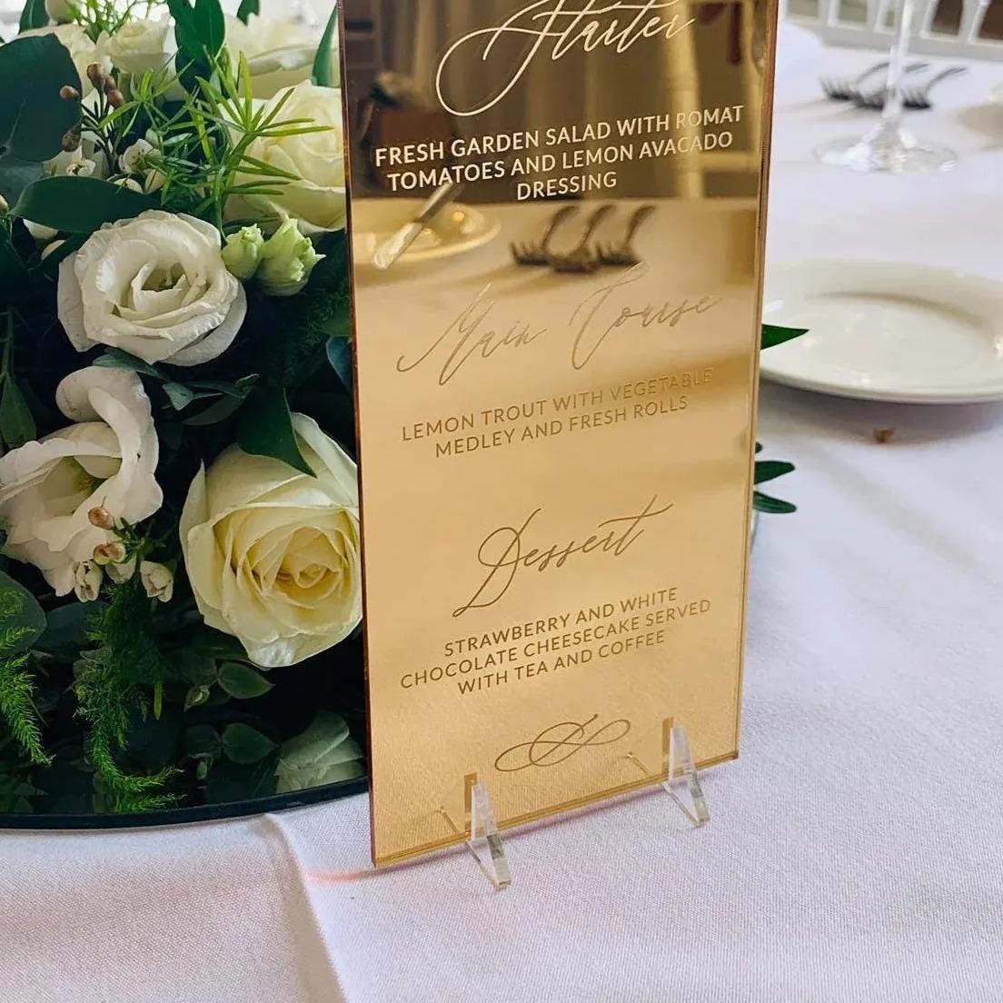 a table setting with a gold menu stand displaying the menu for the starter course. The table is set with a white tablecloth, and there are wine glasses and a vase of white flowers on the table.