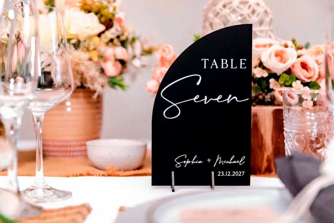 a table setting with a black table sign displaying the number "Seven" and the names "Sophie & Michael" written on it. The table is set with wine glasses, a bowl, and a vase of flowers, creating a festive atmosphere.