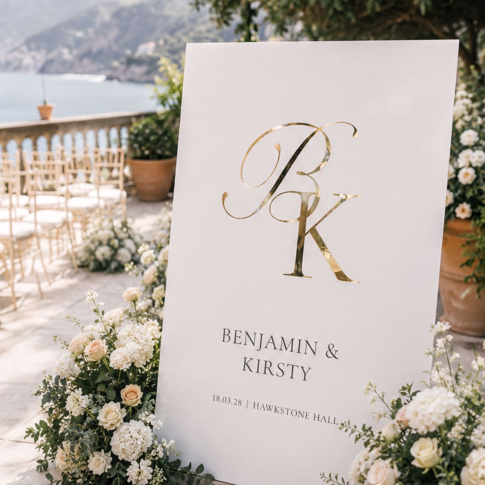 a white sign with a large "B" and "K" in gold lettering, surrounded by white flowers and greenery. The sign displays the names "Benjamin & Kirsty" and the date "18.03.28" at the bottom.