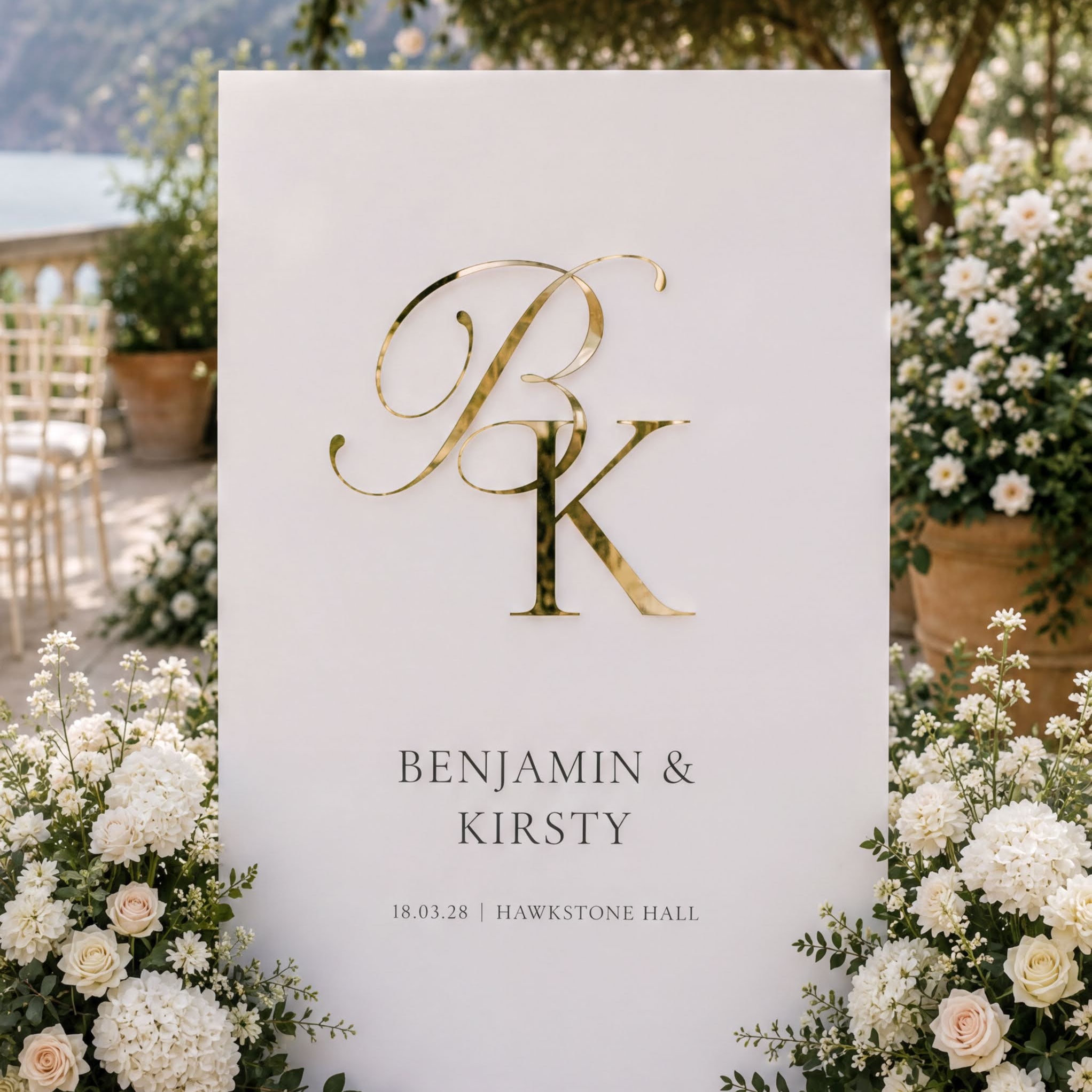 a white sign with a gold letter "P" and the names "BENJAMIN & KIRSTY" written on it, surrounded by white flowers and greenery.
