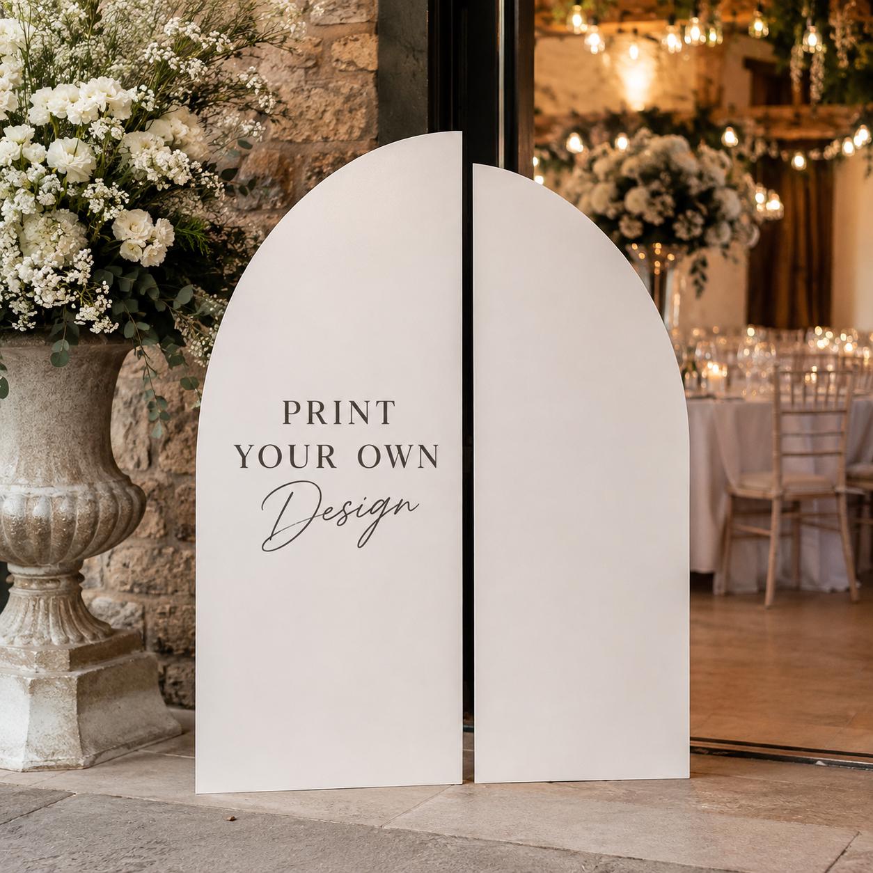 a sign that says "Print Your Own Design" in a white arch shape, placed in front of a building with a stone wall and a large floral arrangement.