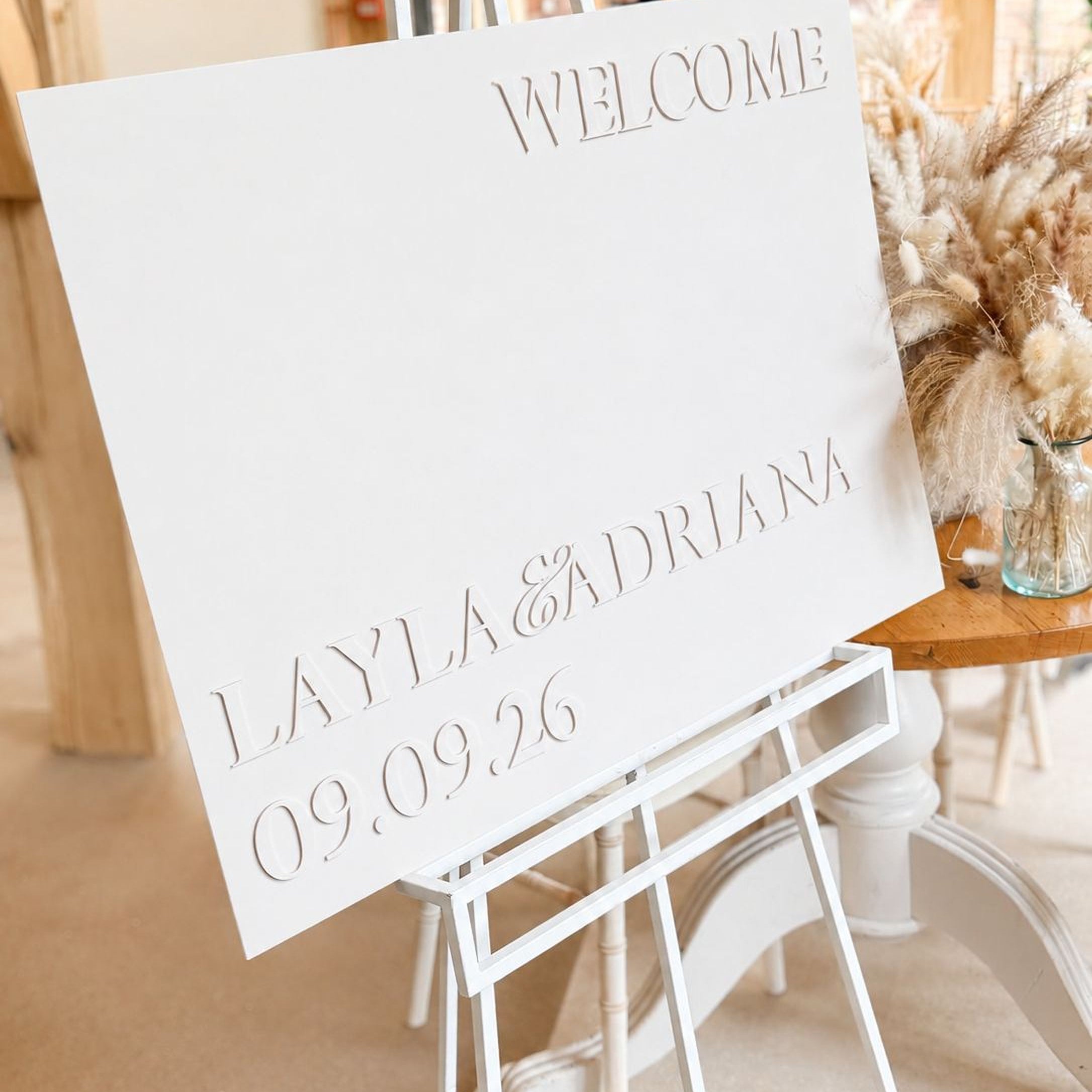 A white sign with the text "WELCOME LAYLA & ADRIANA 09.09.26" is displayed on a white easel.