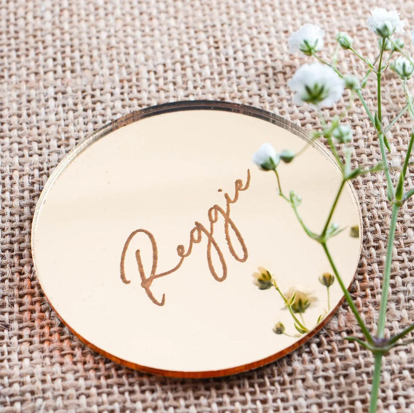 a round, reflective surface with the name "Reggie" written on it in cursive script. The surface is placed on a woven material, and there are small white flowers visible in the background.