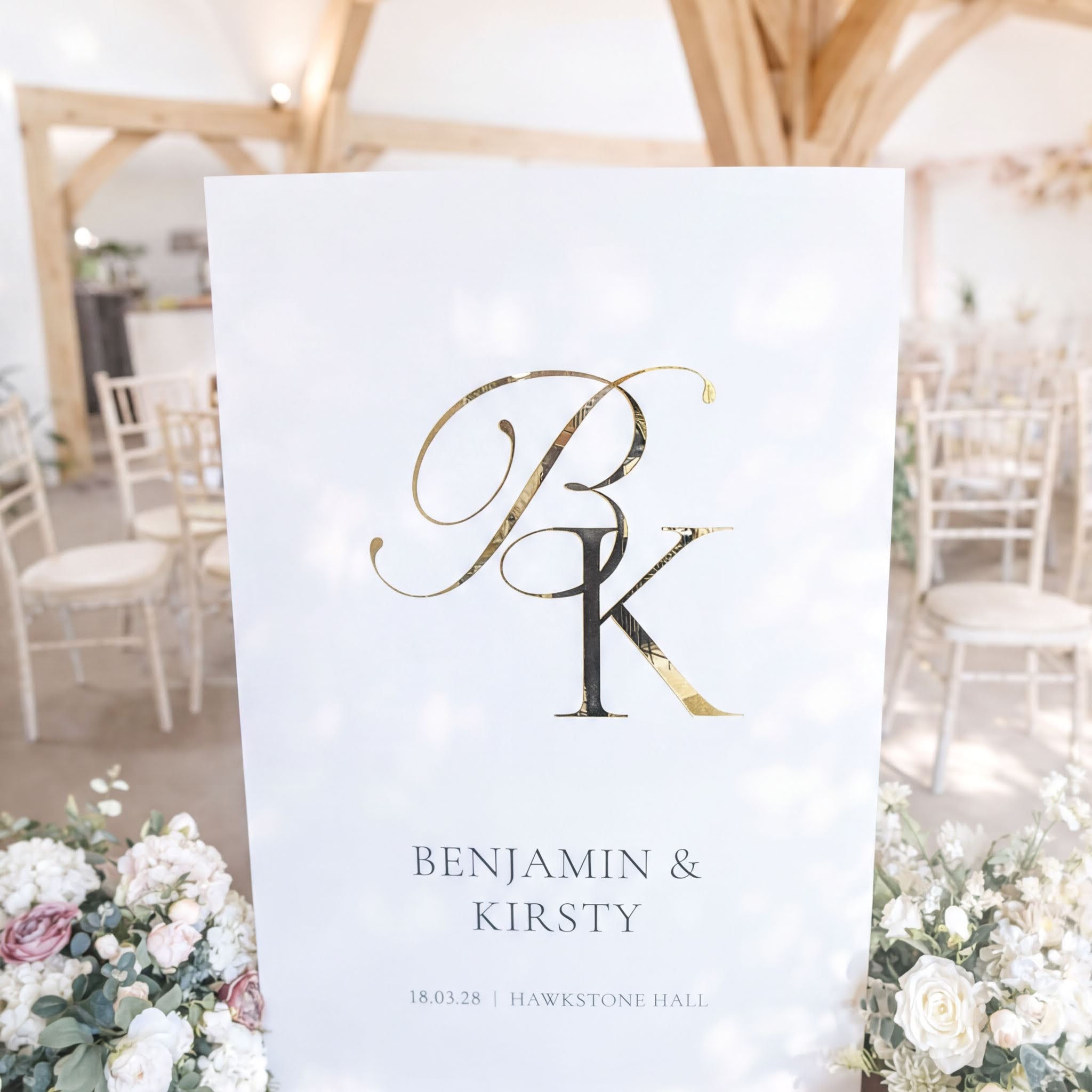 A white sign with the initials "BK" and the names "Benjamin & Kirsty" is displayed in a room with wooden beams and white chairs.