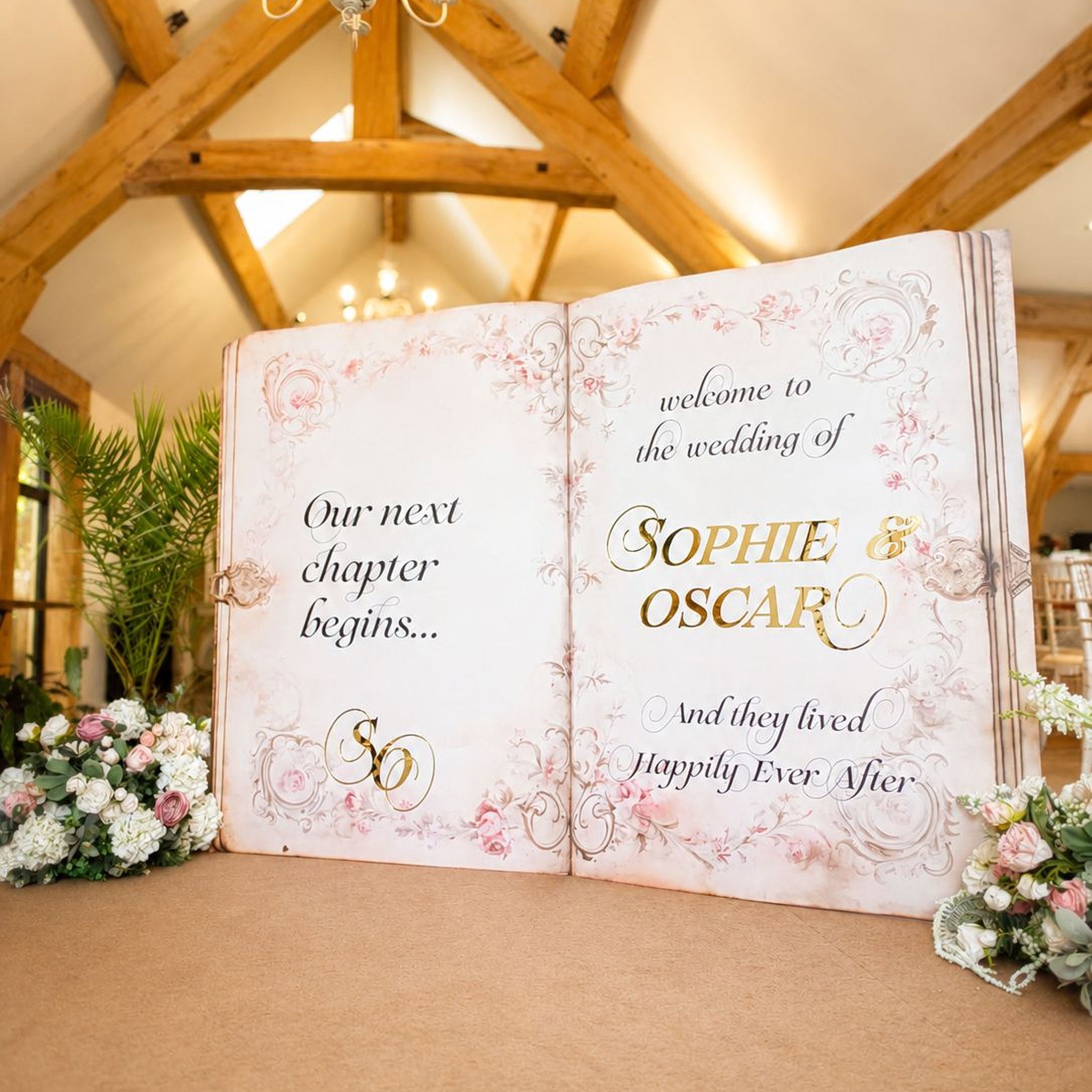 a large open book with a wedding invitation displayed on its pages, set against a wooden background with a chandelier overhead.