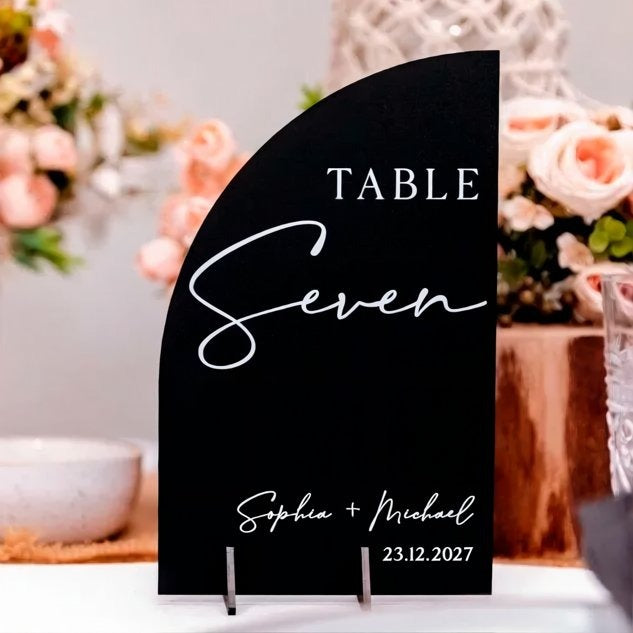 A black table sign with the text "TABLE Seven" and the names "Sophia & Michael" written on it, placed on a table with flowers in the background.