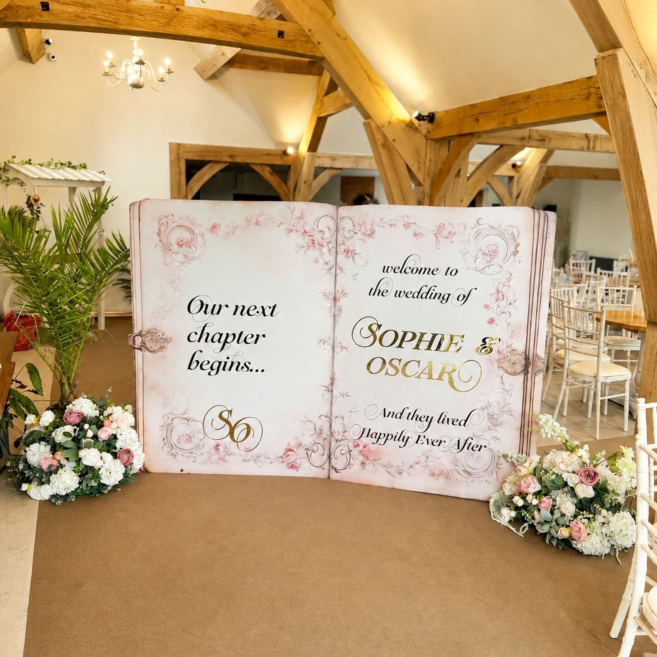 a large open book with a wedding invitation on the cover, surrounded by flowers and a wooden structure in the background.