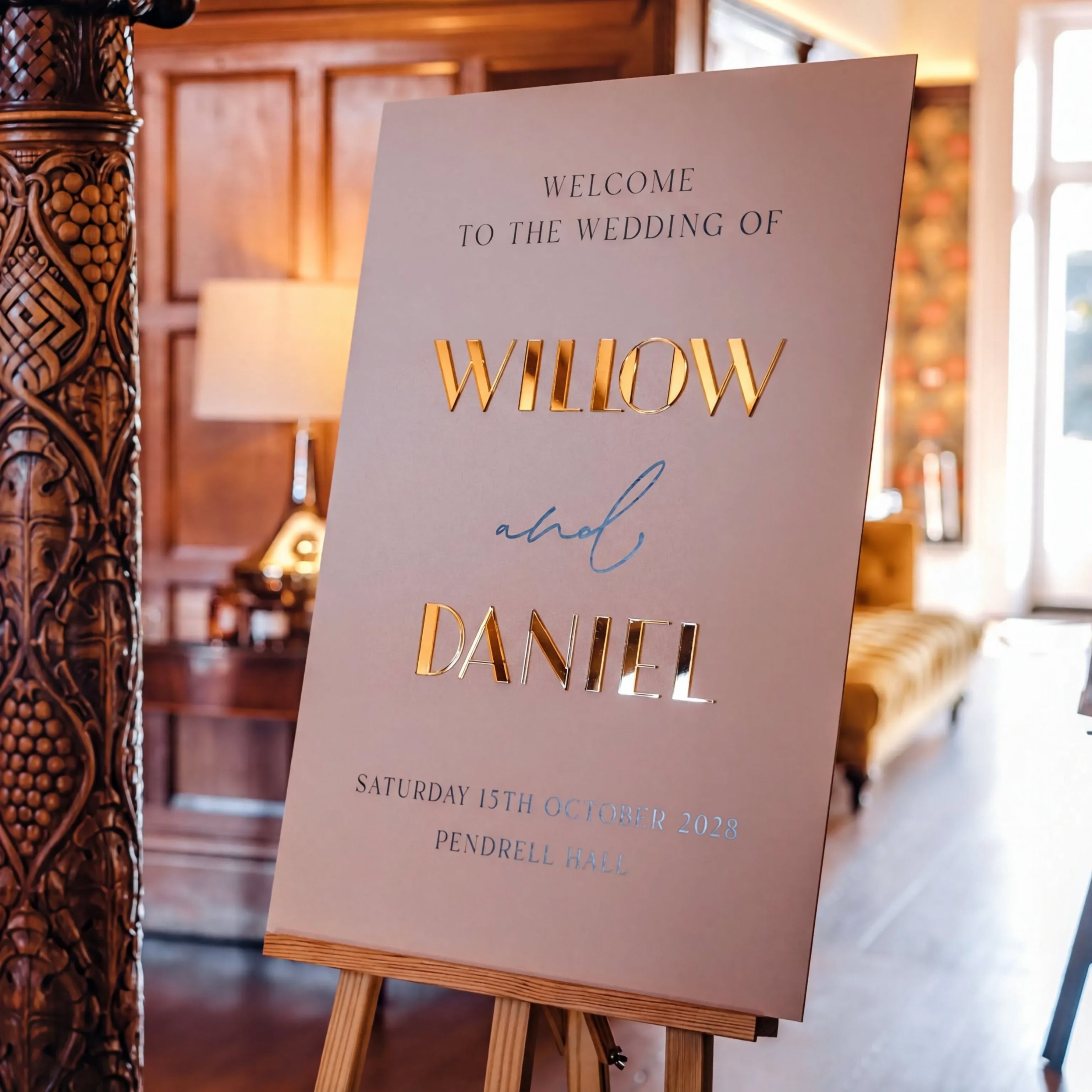 a wedding welcome sign with the names 'WILLOW' and 'DANIEL' prominently displayed. The sign is placed in a room with a wooden paneled wall and a couch, suggesting a formal or elegant setting.