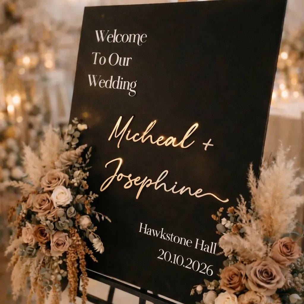 a black sign with gold lettering welcoming guests to a wedding. The sign is decorated with flowers and has the names "Michael + Josephine" written on it. The background of the image is blurred, but it appears to be a wedding reception venue with lights and other decorations.