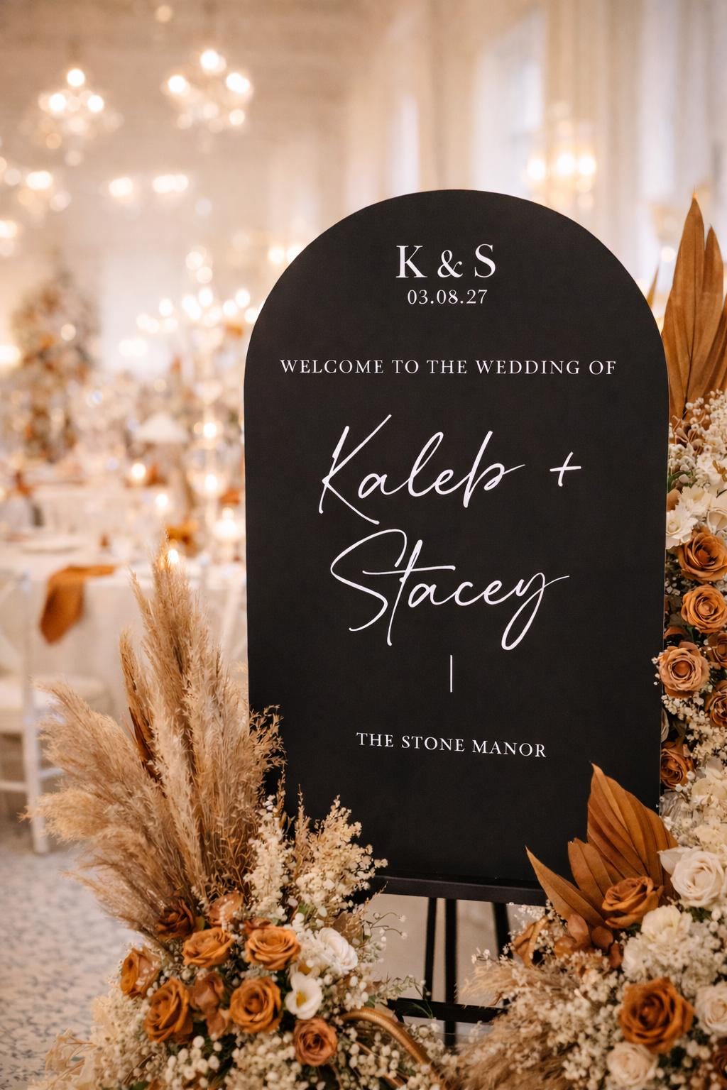 a black sign with white text welcoming guests to the wedding of two people, K&S, on 03.08.27. The sign is surrounded by floral arrangements, including dried flowers and greenery, creating a festive and elegant atmosphere.