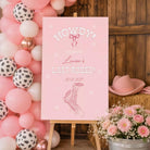 A pink sign welcoming guests to a last rodeo event, with a date and a graphic of a cowboy hat and boots. The sign is set against a backdrop of pink and white balloons, and there is a potted plant and hay bales in the foreground.