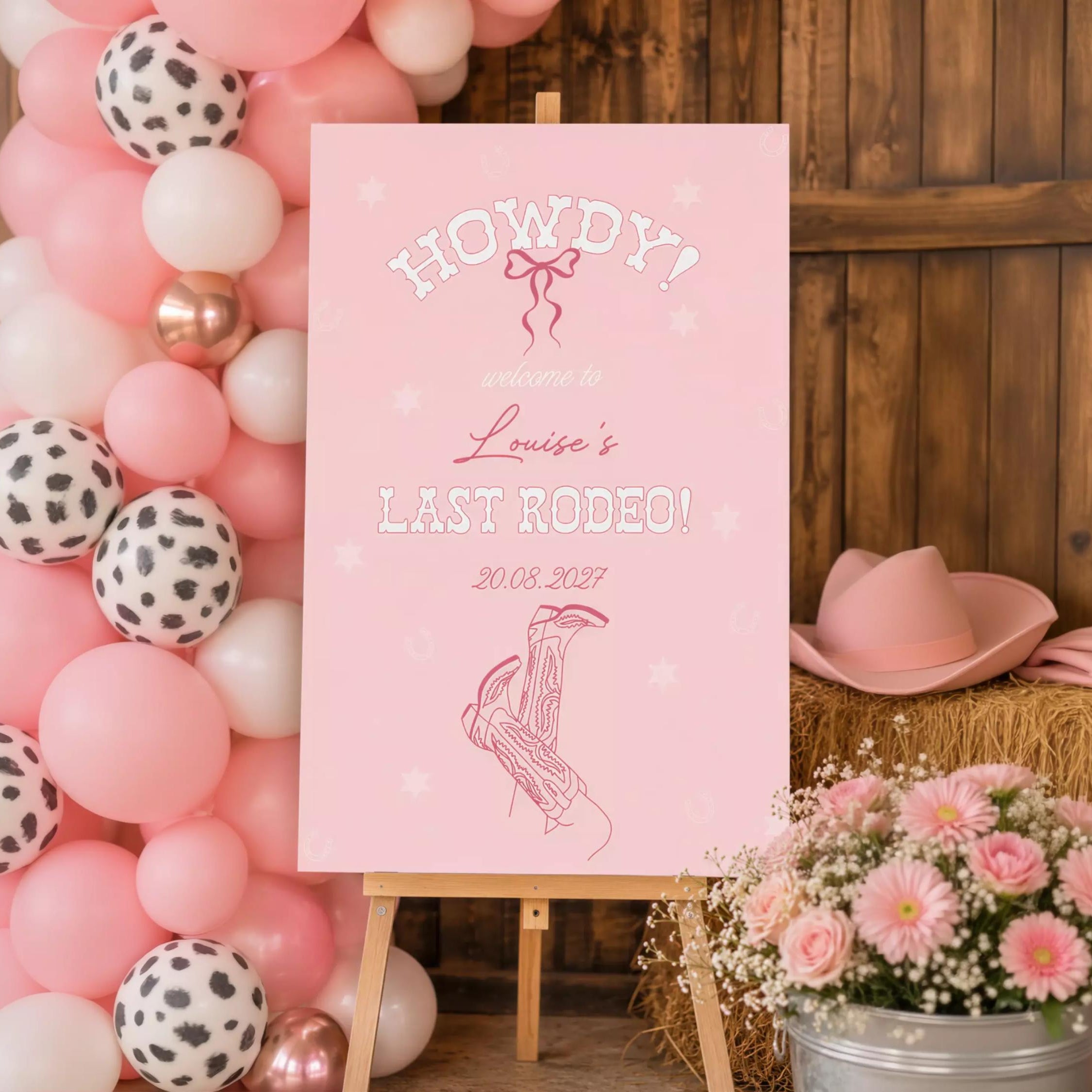 A pink sign welcoming guests to a last rodeo event, with a date and a graphic of a cowboy hat and boots. The sign is set against a backdrop of pink and white balloons, and there is a potted plant and hay bales in the foreground.