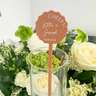A hand holding a small orange card with the text "CHEERS! Katie + Joseph 12.08.2025" against a background of various colorful flowers.