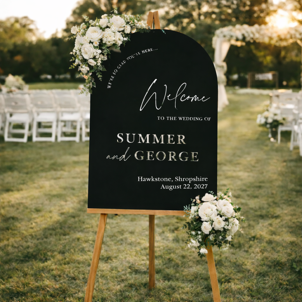 a black sign with white text and floral decorations, standing in a grassy area with trees in the background.