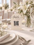 a table setting with a white tablecloth, plates, silverware, and a vase of white flowers. In the foreground, there is a white sign with a QR code and text that reads "Table Eight" and "Scan Here to Pay".