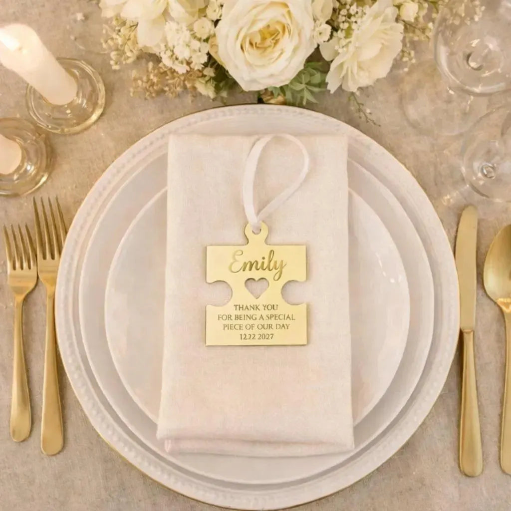 a table setting with a white plate, gold cutlery, and a gold-colored ribbon with a personalized name tag hanging from it. The table is decorated with white flowers and candles, creating a formal and elegant atmosphere.