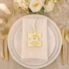 a table setting with a white plate, gold cutlery, and a gold-colored ribbon with a personalized name tag hanging from it. The table is decorated with white flowers and candles, creating a formal and elegant atmosphere.