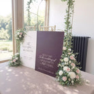 a wedding ceremony setup with two large signs welcoming guests to the wedding of Elizabeth and Daniel. The signs are adorned with white and purple flowers, and the text on the signs reads "Welcome To The Wedding Of Elizabeth And Daniel".