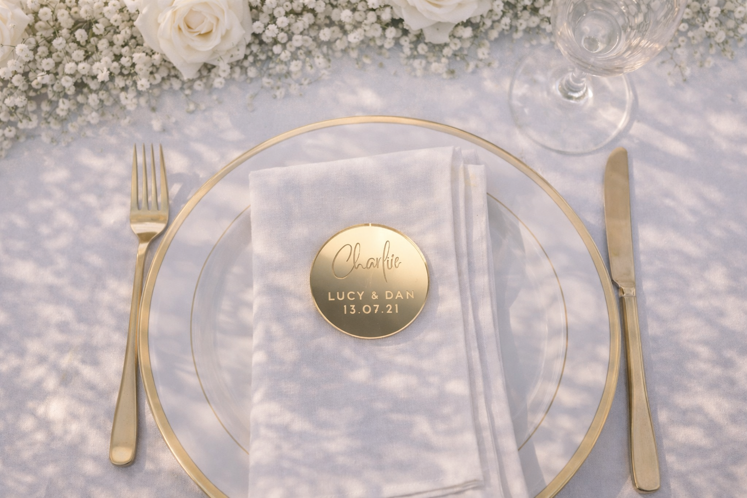 a table setting with a white tablecloth, a gold rimmed plate, a gold fork, a gold knife, and a gold napkin. There is a gold circular plate with a name tag on it, and a glass of wine is also present on the table.
