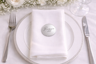 A white place setting with a silver fork, knife, and a round metal name tag on a white napkin.