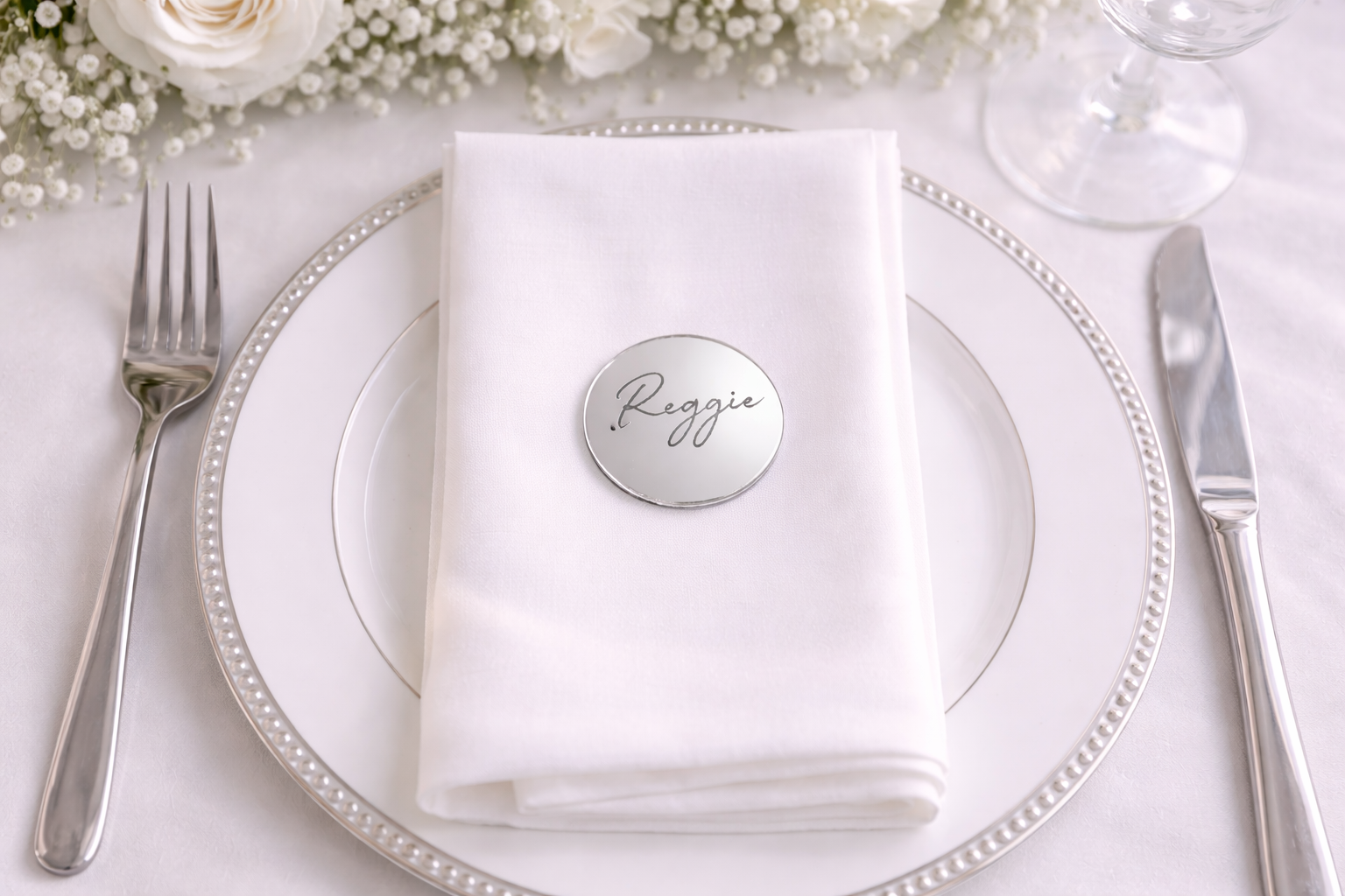 A white place setting with a silver fork, knife, and a round metal name tag on a white napkin.