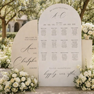 a wedding reception seating plan displayed on a large white sign. The sign is surrounded by white flowers, creating a beautiful and elegant setting.