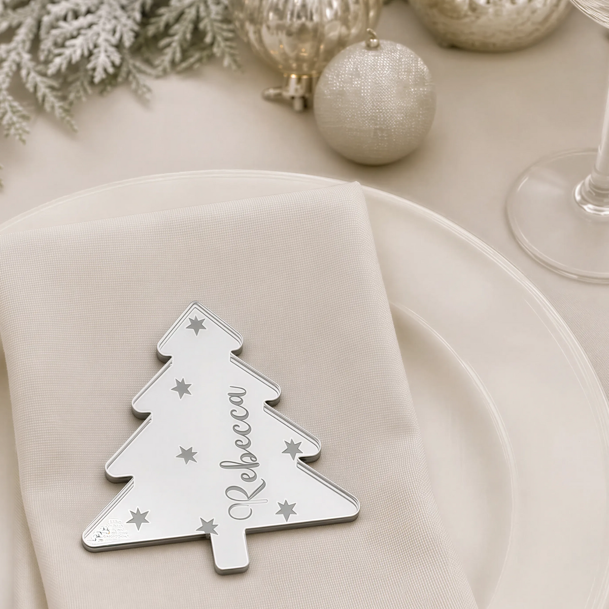 A white plate with a silver tree-shaped nameplate that says "Rebeccah" on it, placed on a table with other Christmas decorations and tableware.