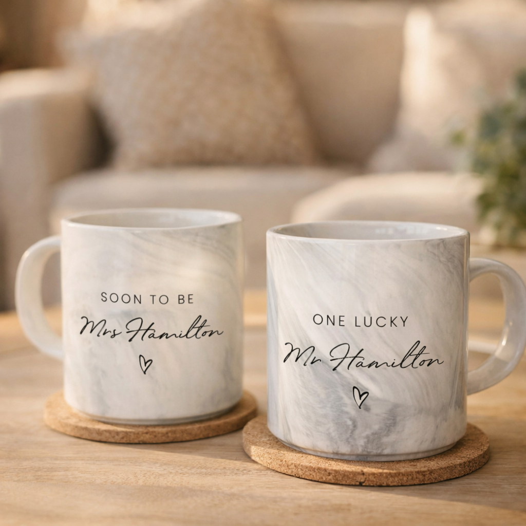 Two white ceramic mugs with text that says "Soon to be Mrs Hamilton" and "One lucky Mr Hamilton" on them, placed on a wooden surface.
