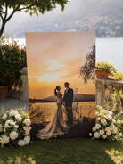 A couple dressed in formal attire stands together in a scenic outdoor setting, with a large framed photograph of them in the foreground and a picturesque landscape in the background.
