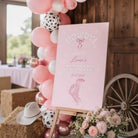 a pink sign with white text and a graphic of a cowboy hat, surrounded by pink and white balloons, a bouquet of pink flowers, and a wooden wagon wheel.