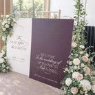 a wedding ceremony setup with two large signs welcoming guests to the wedding of Elizabeth and Daniel. The signs are adorned with white and purple flowers, and the text on the signs reads "Welcome To The Wedding Of Elizabeth And Daniel".