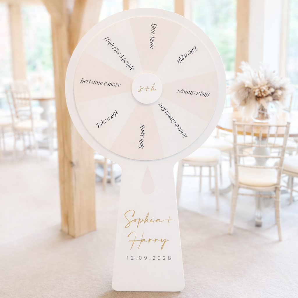 A white spinning wheel with gold text and a gold name tag, standing in a room with a wooden beam and chairs in the background.