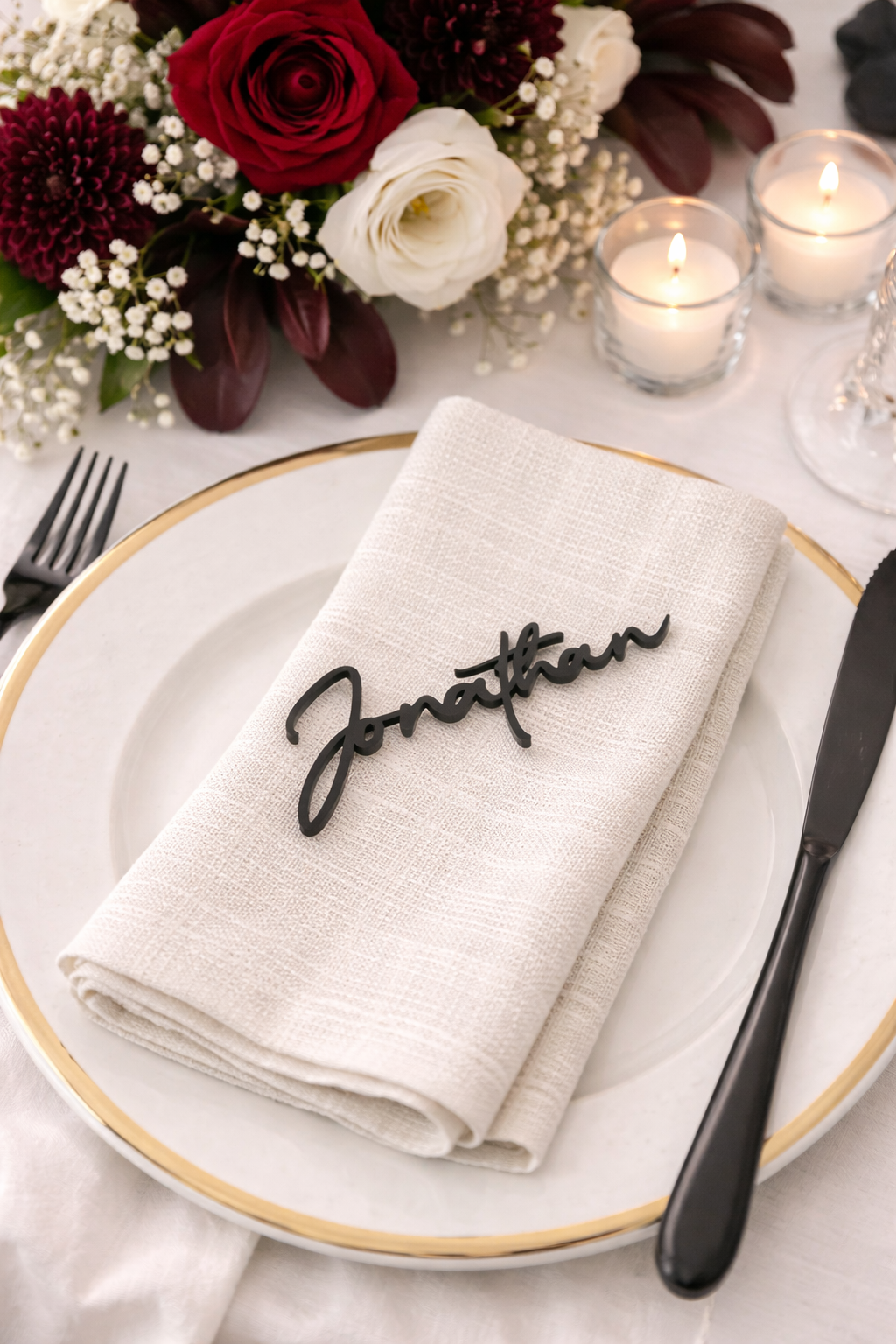 a table setting with a white plate, a folded napkin with a name tag, and a vase of red and white flowers. There are also lit candles on the table.
