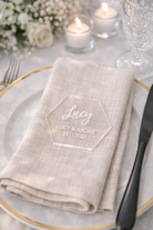 A table setting with a white napkin, a glass of water, and a candle, with a clear hexagonal placemat displaying the name 'Lucy & Archie' and the date '23.11.2023'.
