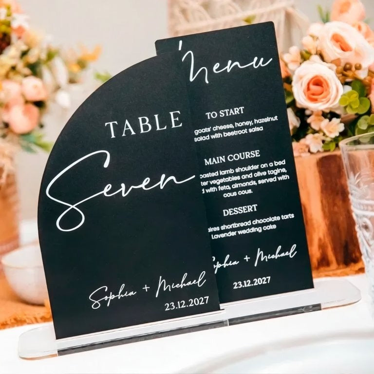 a black table menu with white text, placed on a table with a vase of flowers in the background.