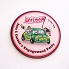 A round coaster with a cartoon illustration of a couple in a green car, celebrating an engagement party.