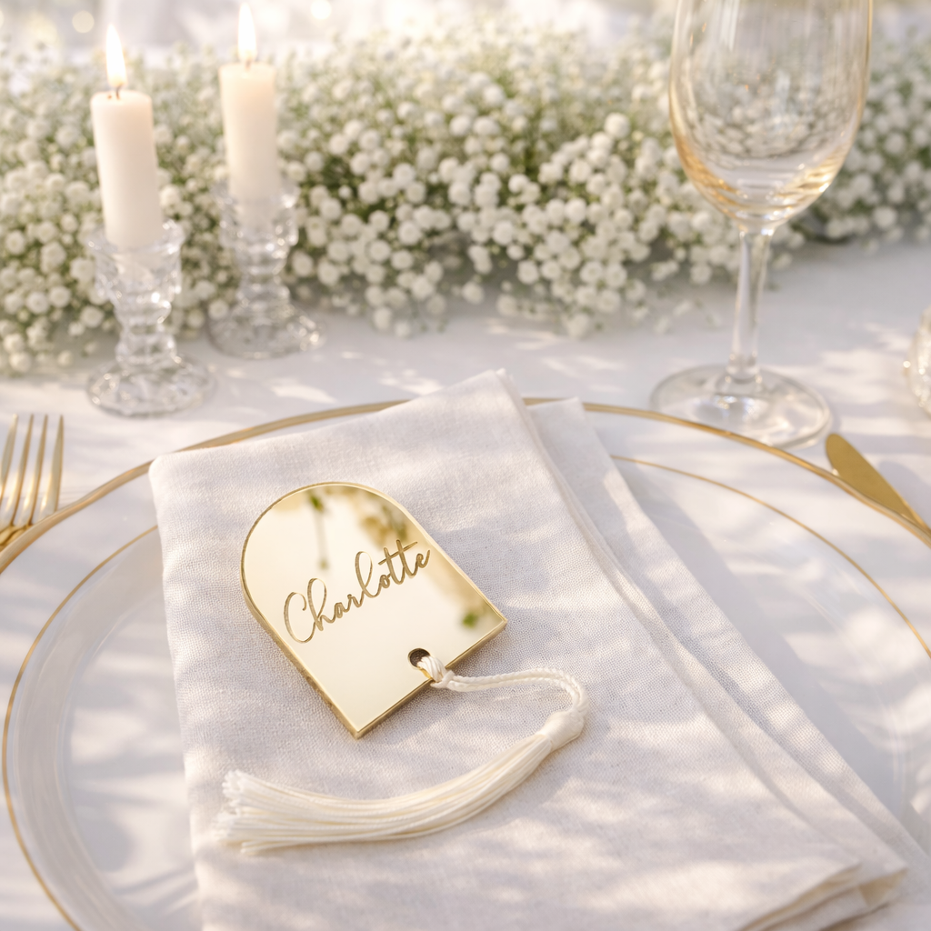 a table setting with a white tablecloth, a gold-rimmed plate, and a white napkin with a name tag. There are also two candles and a wine glass on the table.
