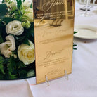 a table setting with a gold menu stand displaying the menu for the starter course. The table is set with a white tablecloth, and there are wine glasses and a vase of white flowers on the table.